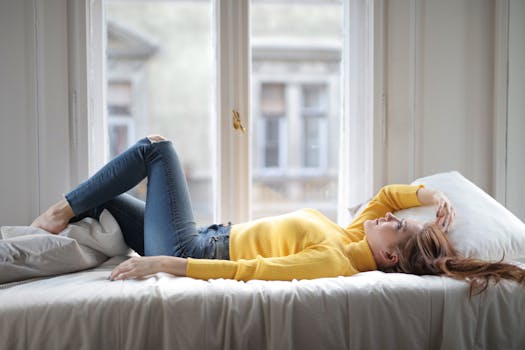 A young woman in a yellow sweater relaxes on a bed by a window, embodying tranquility and comfort.
