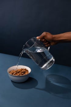 A person pouring water from a glass pitcher into a bowl of almonds on a blue surface.