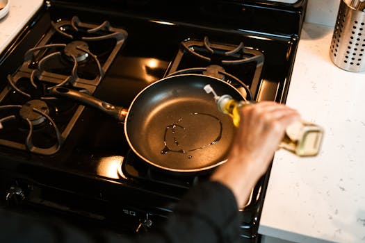 From above crop faceless chef pouring olive oil into frying pan on stove while cooking at home