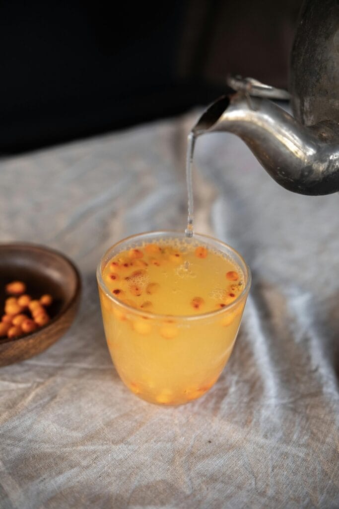 A warm sea buckthorn tea being poured from a kettle into a glass on a linen tablecloth.