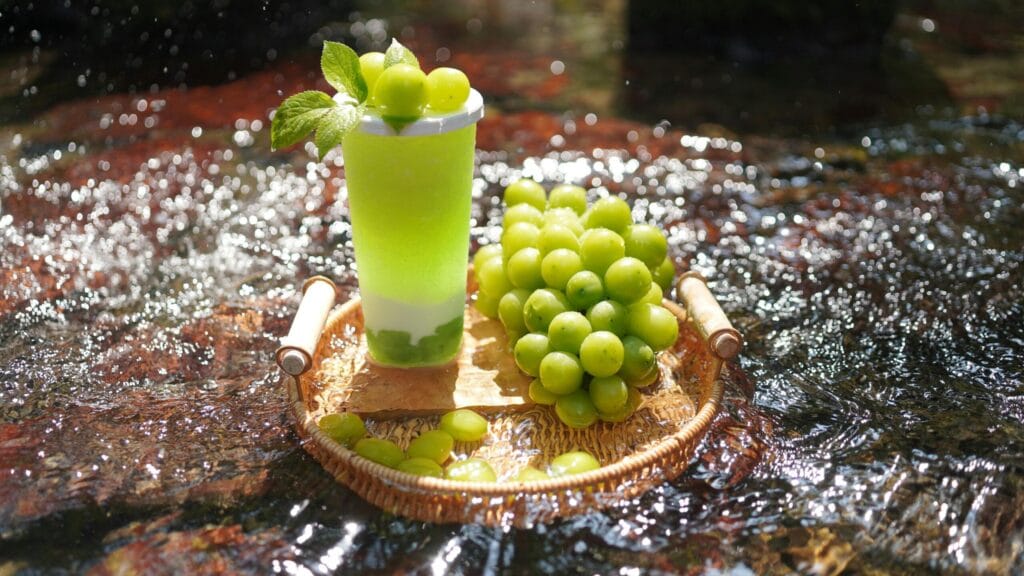 A green grape drink set beside fresh grapes on a wicker tray in a natural outdoor stream.