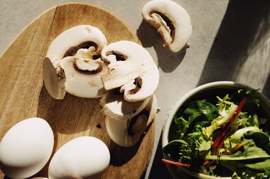 Sliced mushrooms with eggs on a wooden board beside fresh salad greens.