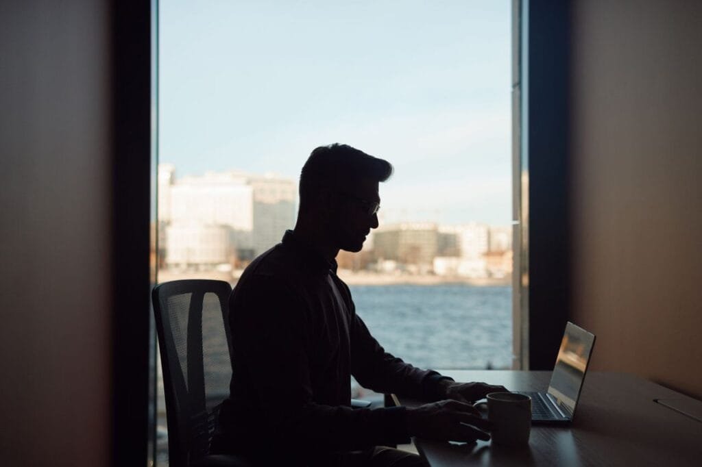 Silhouette of a man working on his laptop by a window overlooking a waterfront cityscape.