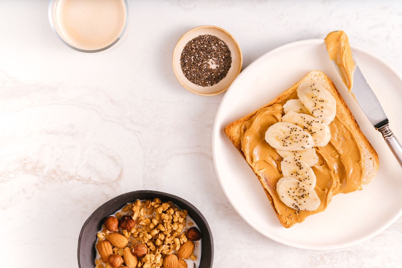 Flat lay of a nutritious breakfast featuring banana toast, chia seeds, and nuts on a marble surface.